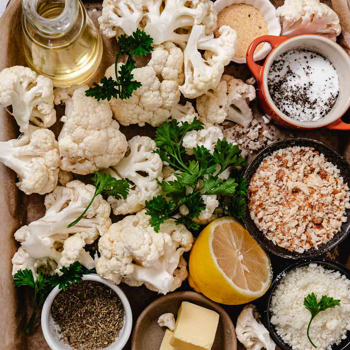 Raw cauliflower florets with olive oil, butter, lemon, fresh parsley, breadcrumbs, herbs, salt, pepper, and grated cheese arranged on a baking tray
