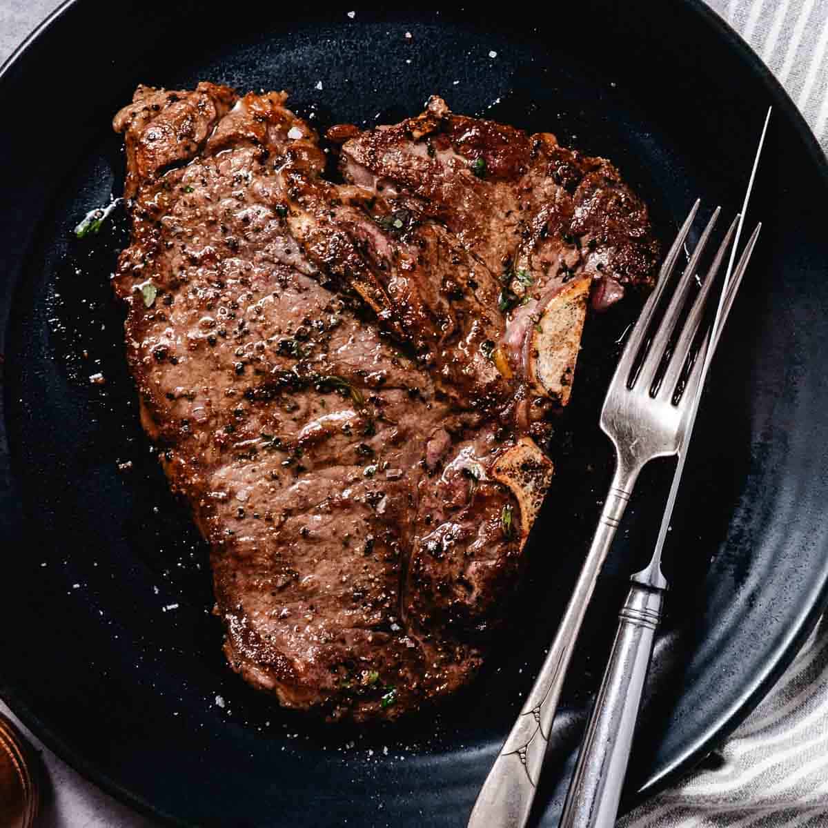 Cooked t-bone steak on a dark plate with knife and fork.