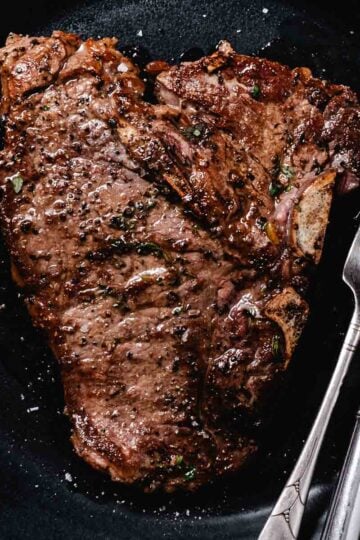 Cooked thin t-bone steak on a dark plate with knife and fork on the right side.