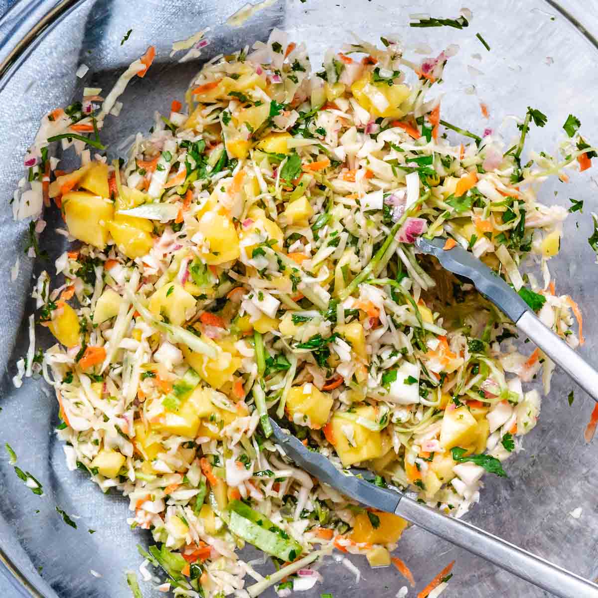 Mango coleslaw being tossed in a glass bowl with cabbage, cilantro, carrot, and red onion.
