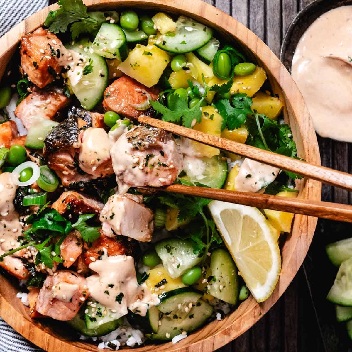 Marinated salmon rice bowl with seared salmon cubes, white rice, cucumber, edamame, mango, cilantro, sesame seeds, and creamy sauce, served with chopsticks and a lemon wedge.