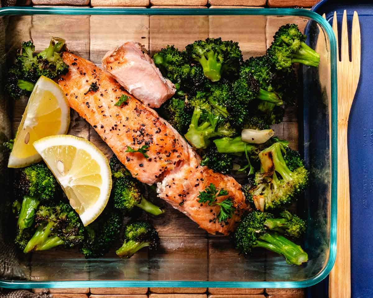 Lemon garlic salmon with roasted broccoli stored in a glass meal prep container with lemon wedge.