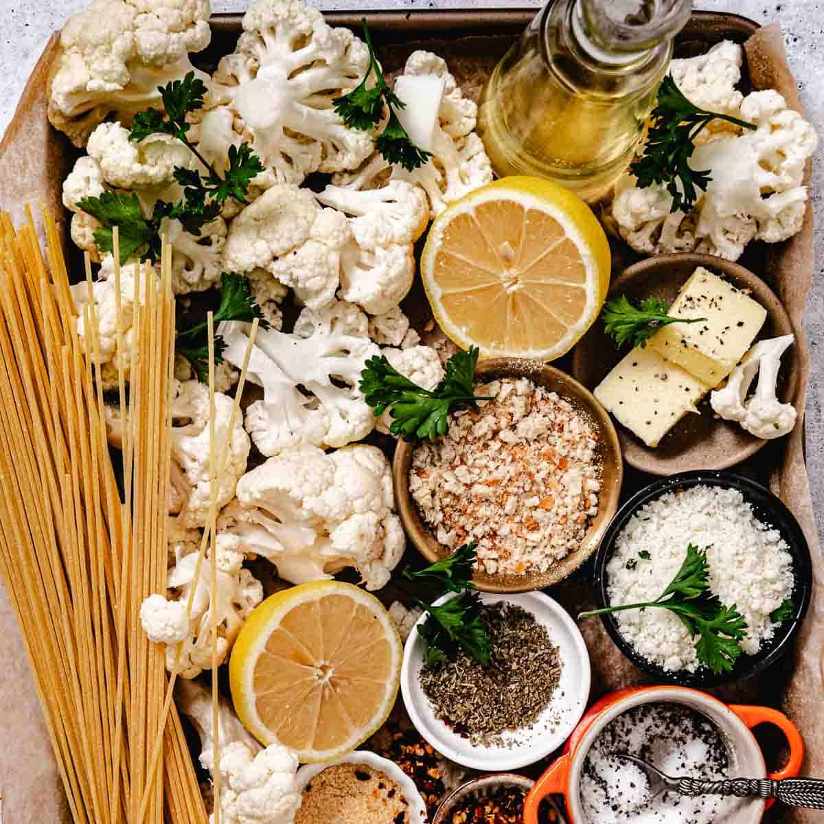 Overhead shot of raw cauliflower florets, uncooked spaghetti, halved lemons, olive oil, butter, breadcrumbs, Parmesan cheese, herbs, salt, pepper, and seasonings arranged on a tray before cooking.