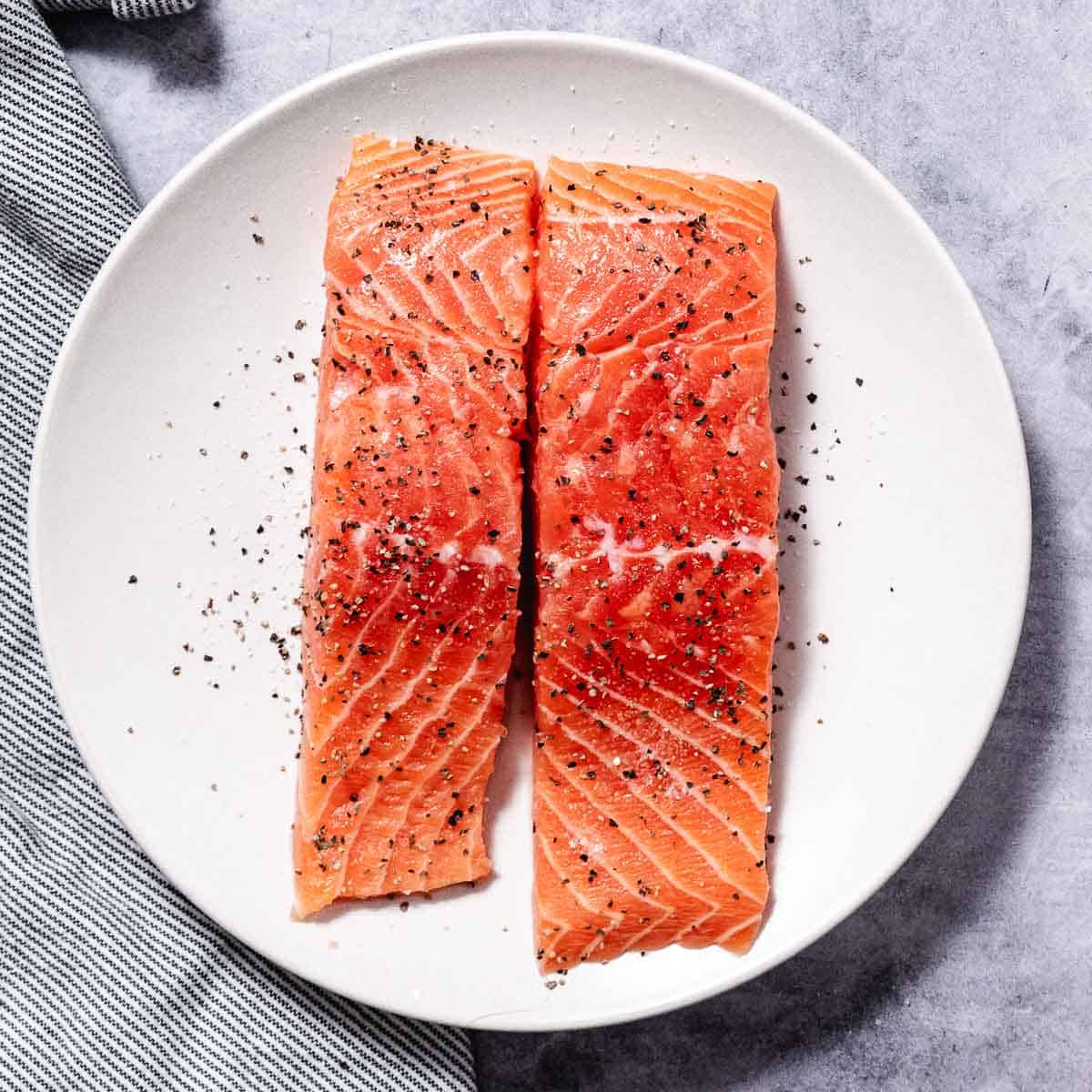 Raw skinless salmon fillets seasoned with salt and black pepper on a white plate.