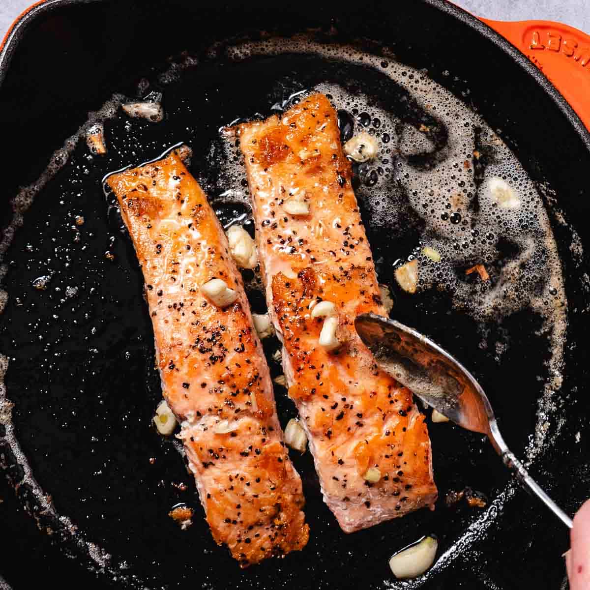 Skinless salmon fillets searing in a cast iron skillet with butter and sliced garlic.