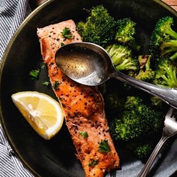 Lemon garlic seared salmon with roasted broccoli in a skillet, topped with fresh parsley and served with a lemon wedge while butter is spooned over the fish.