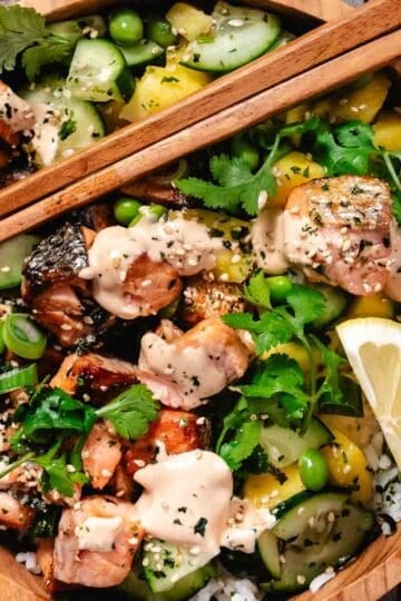 Marinated salmon rice bowl topped with cucumber, mango, edamame, cilantro, furikake, and creamy sriracha mayo, served with chopsticks and a lemon wedge.