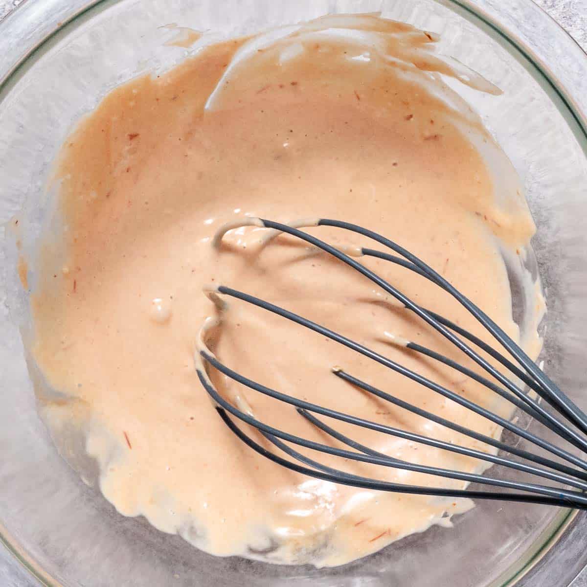 Creamy sriracha mayo dressing being whisked in a clear glass bowl, smooth and lightly speckled with seasoning.