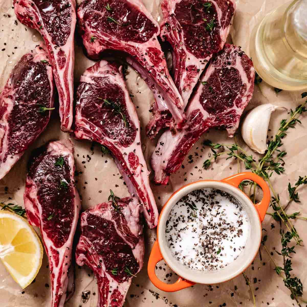 Raw lamb chops arranged on parchment paper with olive oil, garlic, thyme, salt, pepper, and a lemon wedge.