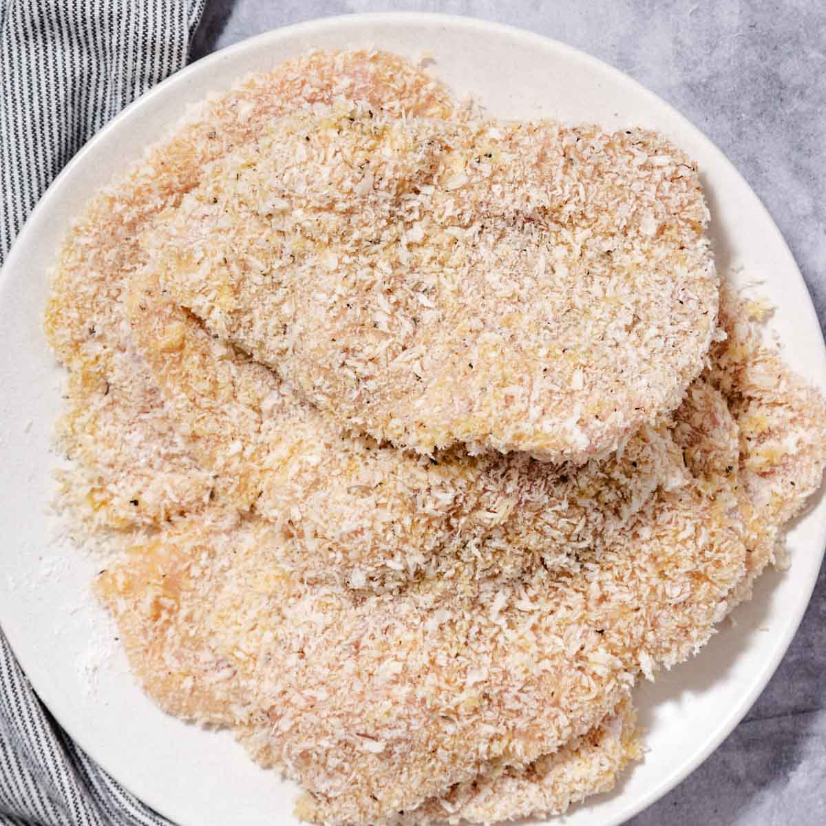 Raw chicken cutlets evenly coated in a thick layer of panko breadcrumbs and herbs on a white plate, ready for pan-frying.