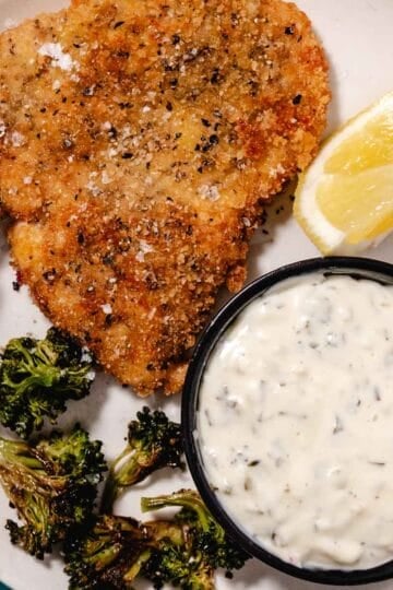 Fried breaded swordfish steak with lemon wedge, roasted broccoli, and a bowl of tartar sauce on a dinner plate.