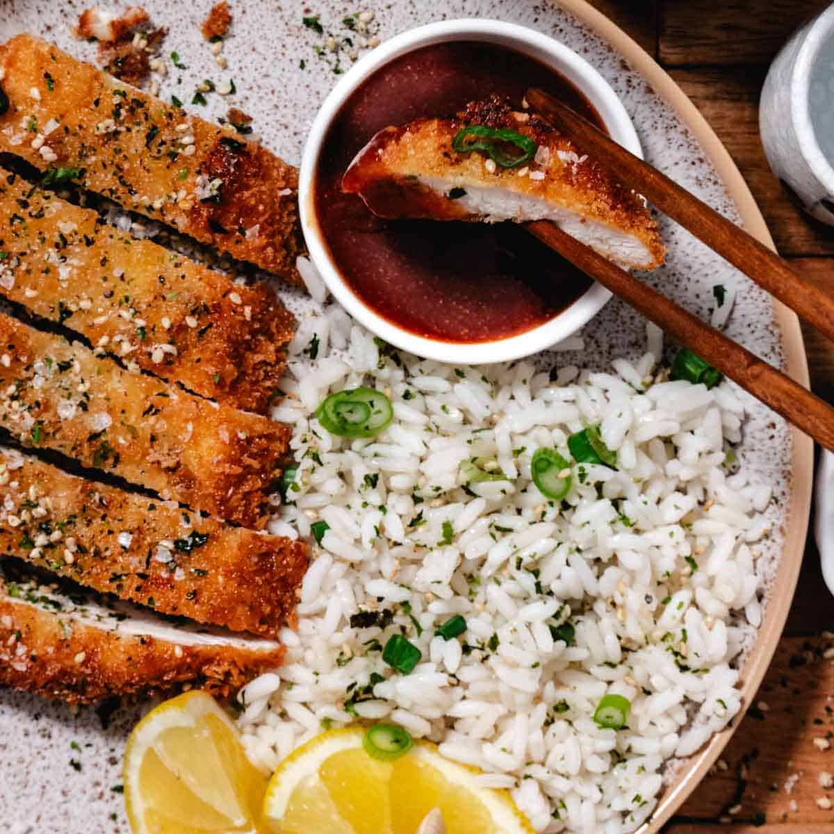 Chicken katsu dipped in sauce with chopsticks, served with white rice, sesame seeds, and lemon slices on a plate