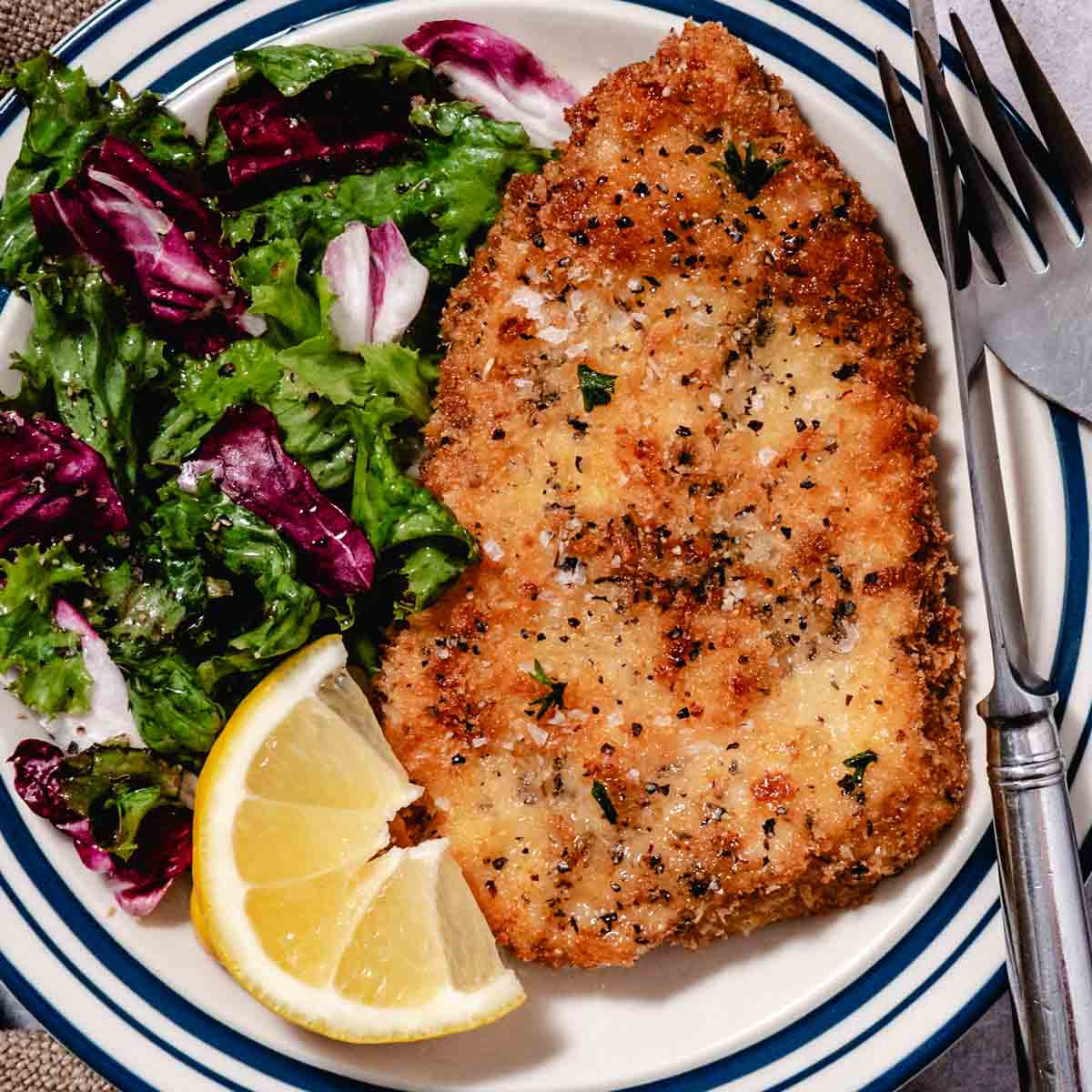 Plated Panko Chicken Milanese with a crispy golden crust and flaky sea salt, served with market-fresh mixed greens, radicchio, and a bright lemon wedge on a blue-rimmed plate.