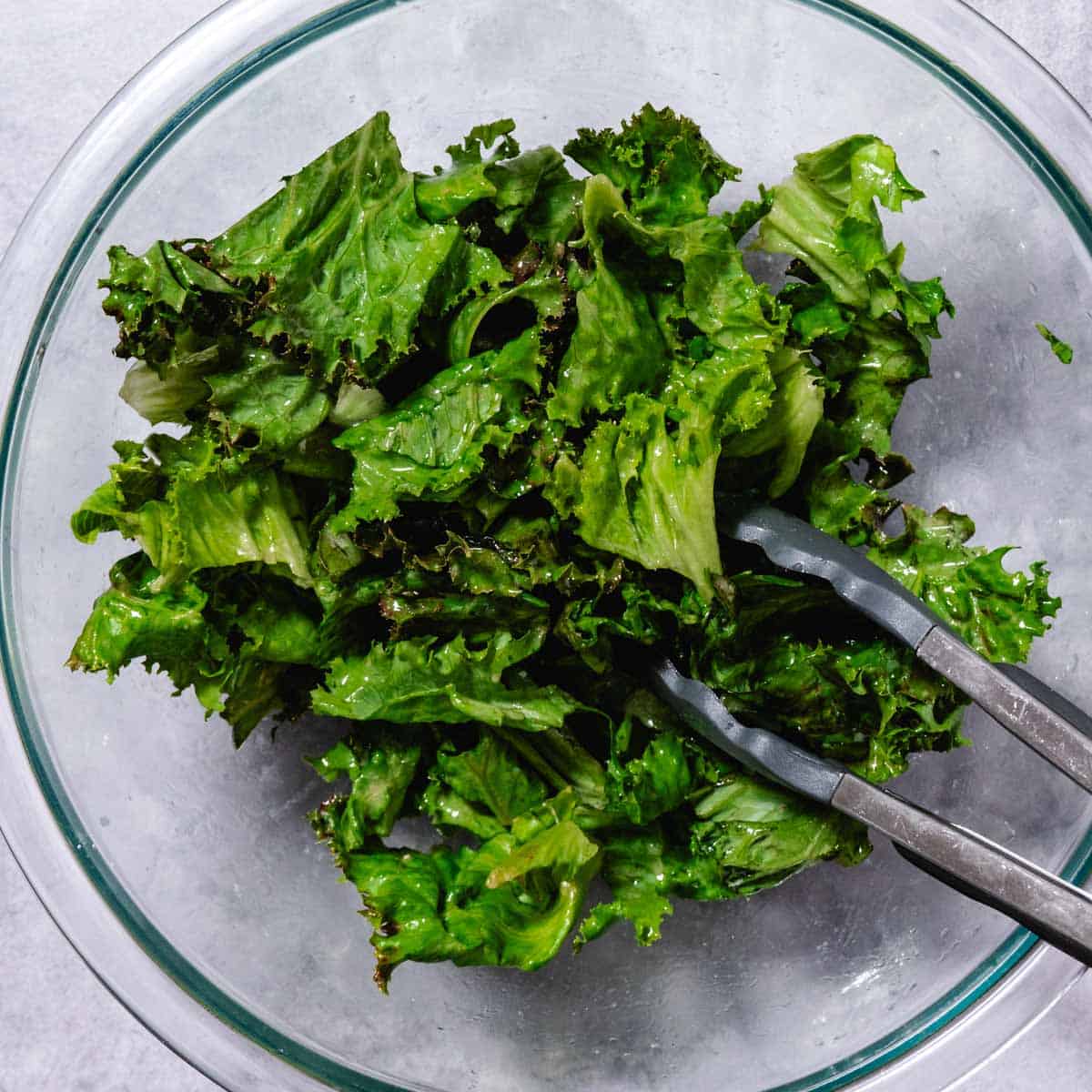 A glass bowl of market-fresh mixed greens, including ruffled green leaf lettuce and purple radicchio, lightly tossed in a translucent red wine vinaigrette with silver serving tongs.