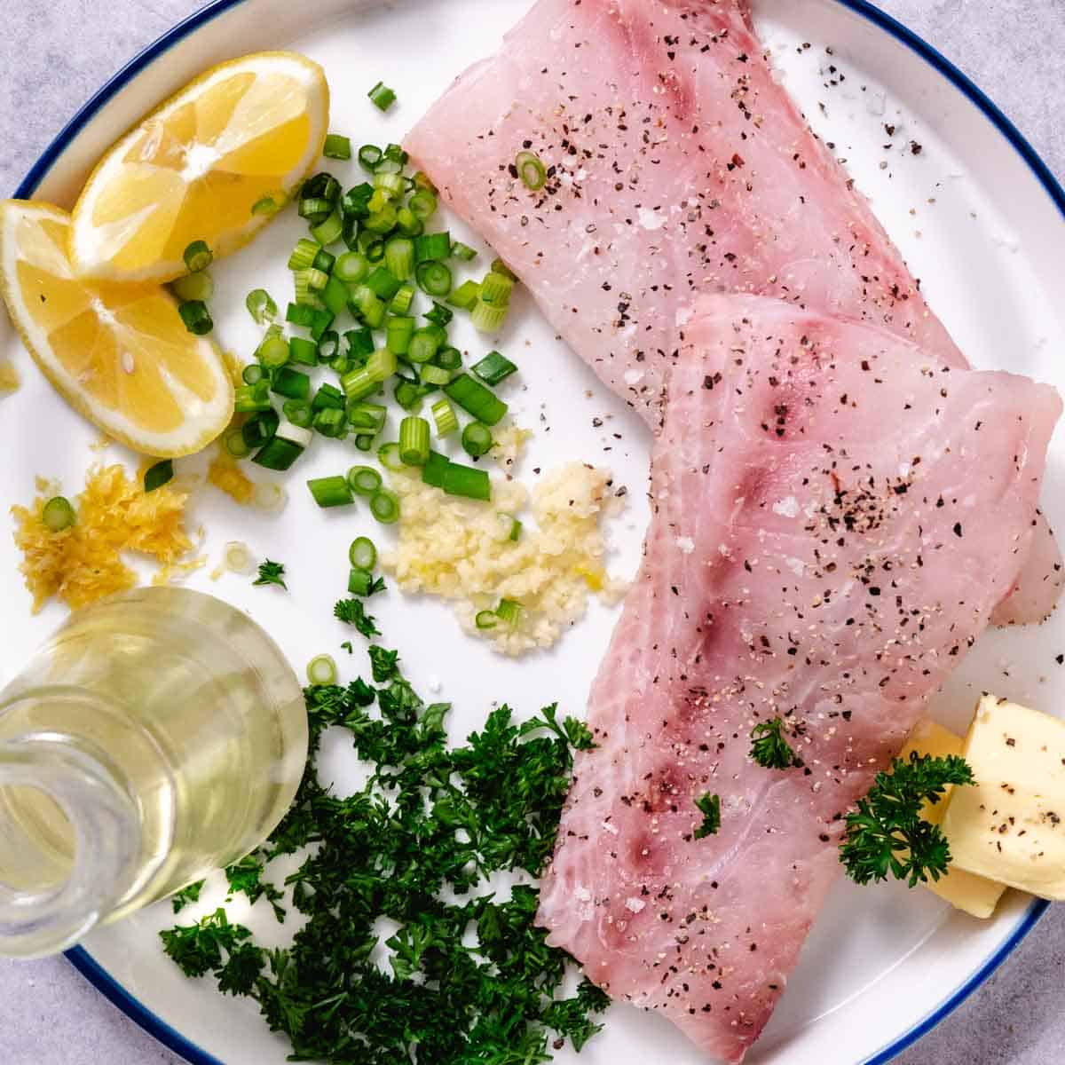 Raw tilefish fillets with lemon, butter, garlic, parsley, green onions, and salt and pepper on a cutting board.