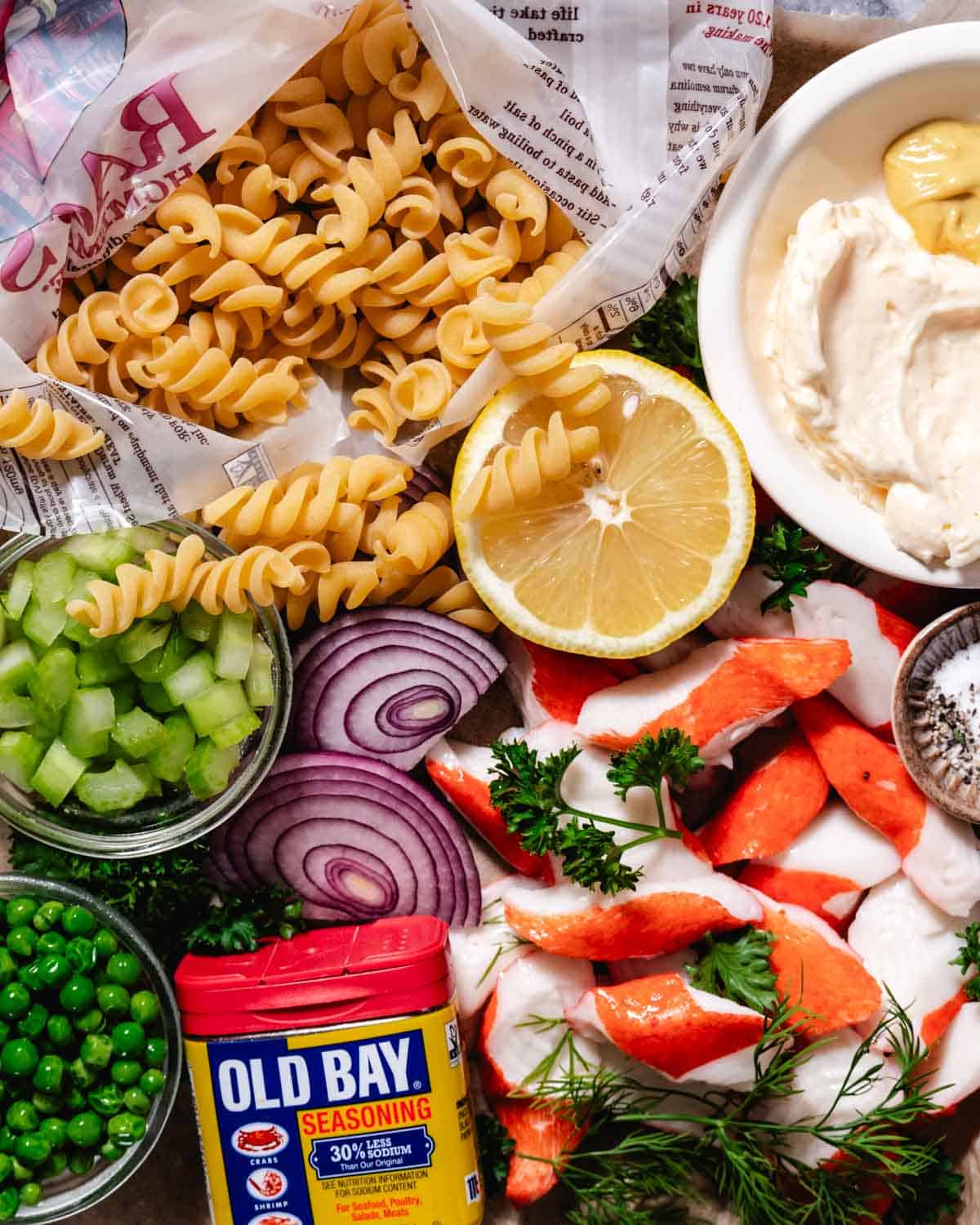 Ingredients for crab meat pasta salad including fusilli pasta, imitation crab, celery, red onion, peas, lemon, mayonnaise, Dijon, and Old Bay seasoning.