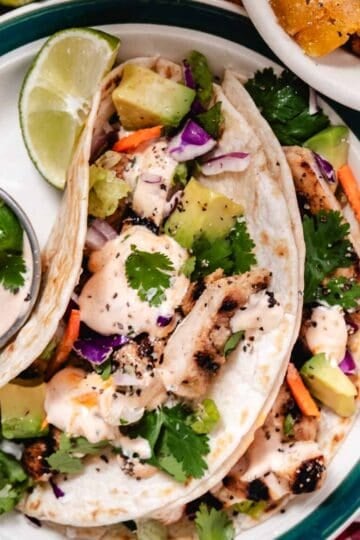 Grilled chicken tacos with avocado, cabbage slaw, cilantro, and creamy taco sauce served with lime wedges and dipping sauce.
