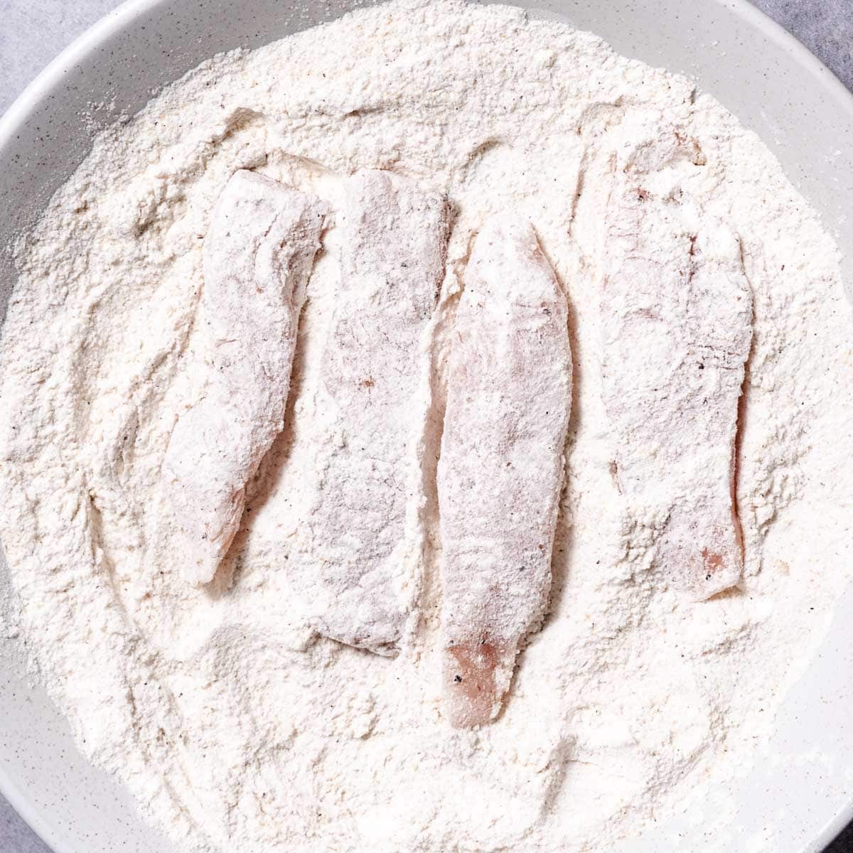 Chicken strips coated in seasoned flour in a shallow bowl.