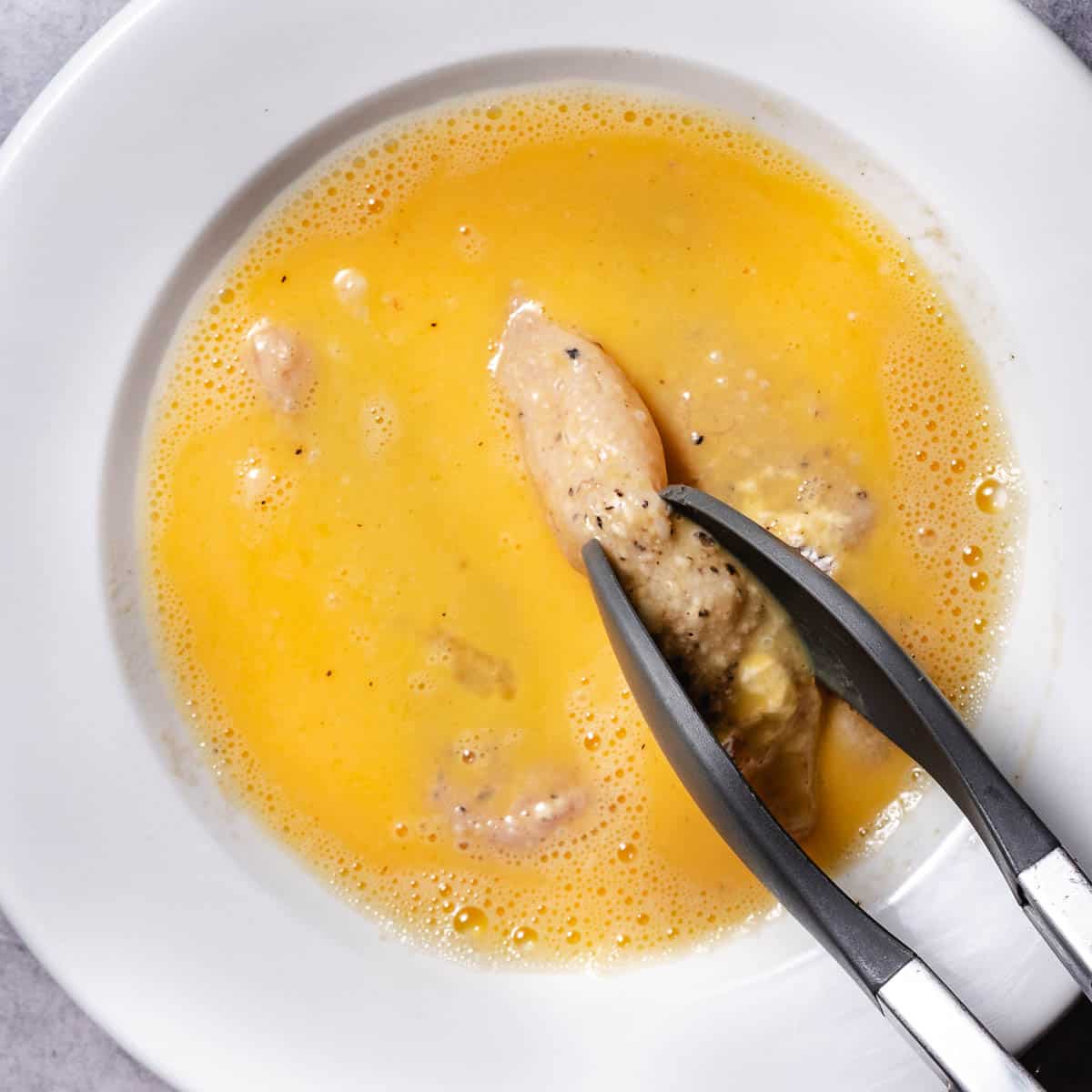 Floured chicken strip dipped into beaten egg using kitchen tongs.