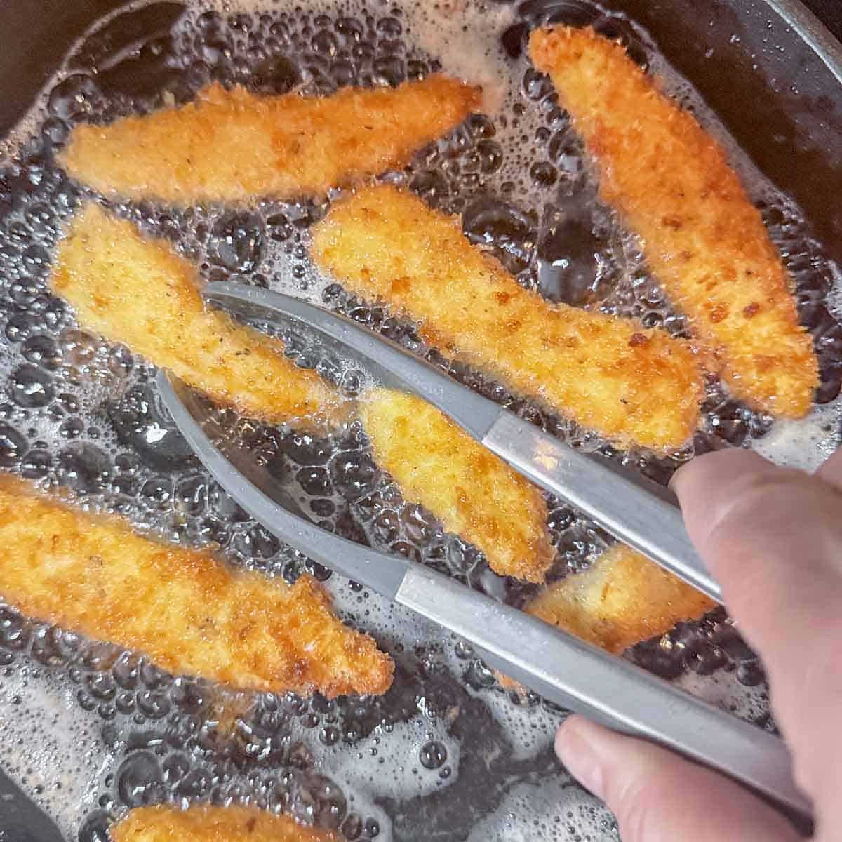 Panko-breaded chicken strips frying in hot oil and being turned with tongs.