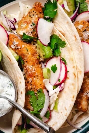 Crispy chicken tacos with panko-breaded chicken, shredded cabbage, avocado, radishes, and red onion in soft tortillas, topped with a creamy herb sauce and served with lime wedges.