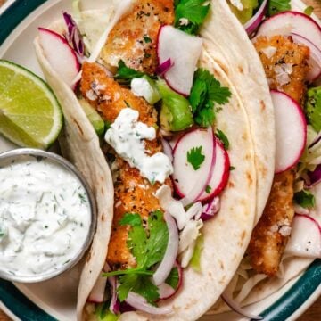 Crispy chicken tacos with panko-breaded chicken, shredded cabbage, avocado, radishes, and red onion in soft tortillas, topped with a creamy herb sauce and served with lime wedges.