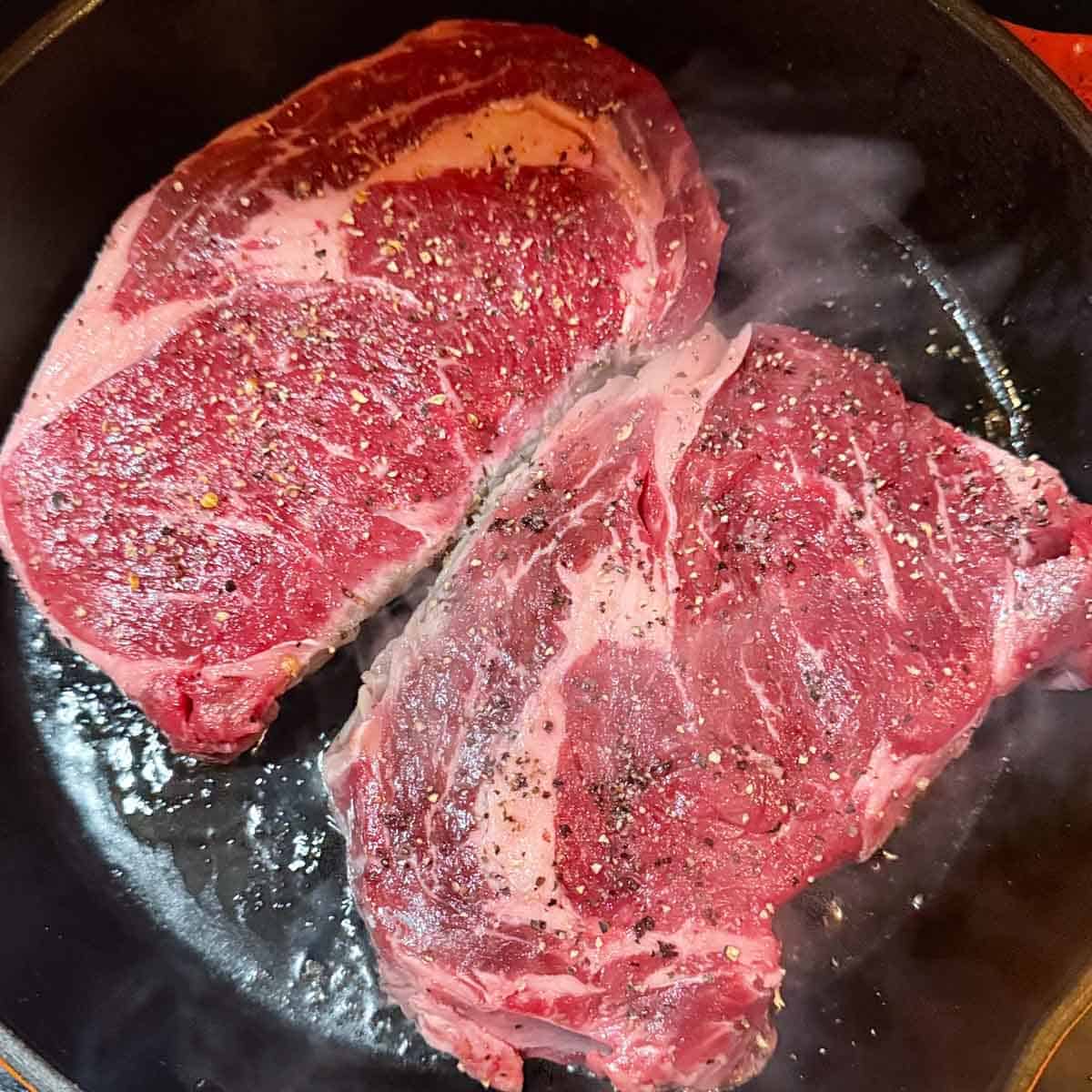 Raw ribeye steaks seasoned with salt and pepper in a hot cast iron skillet