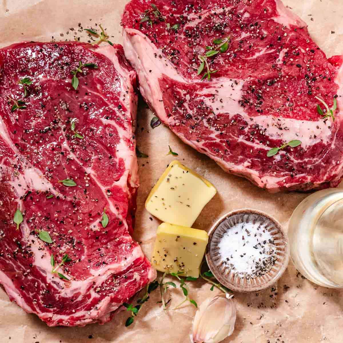 Raw ribeye steaks with butter, garlic, salt, and pepper on parchment paper