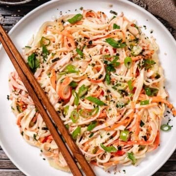 Kani salad on a white plate with shredded imitation crab, cucumber, carrots, green onions, and sesame seeds, served with chopsticks.