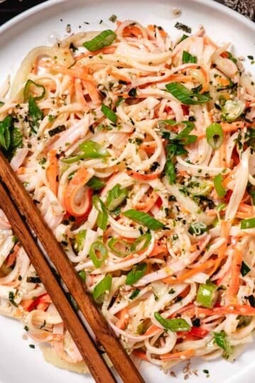 Kani salad on a white plate with shredded imitation crab, cucumber, carrots, green onions, and sesame seeds, served with chopsticks.