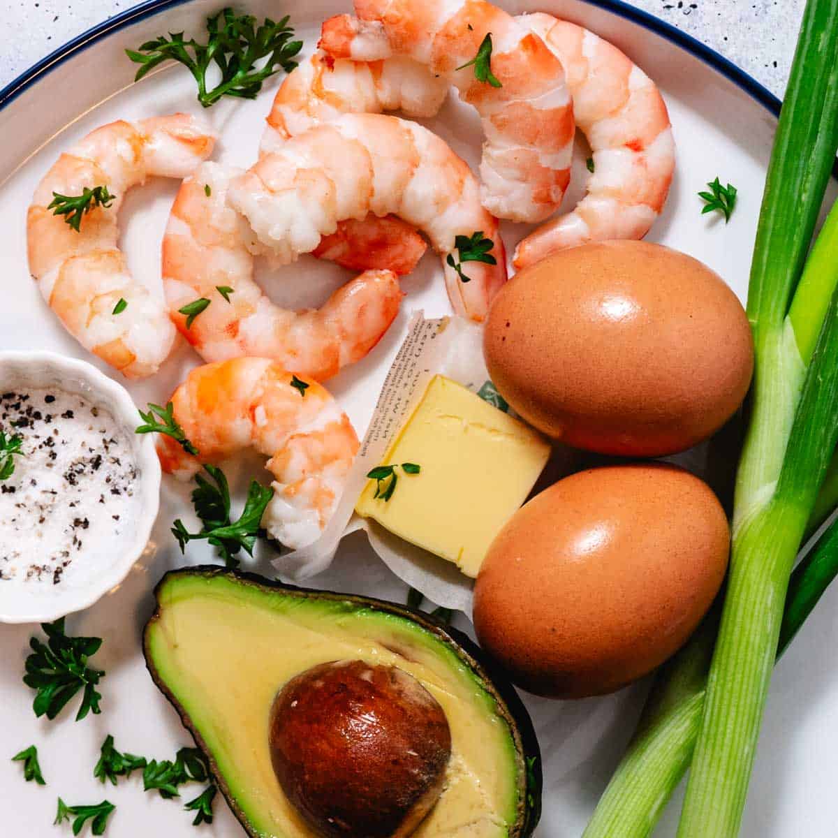 Ingredients for shrimp and scrambled eggs including cooked shrimp, eggs, butter, scallions, avocado, salt, and pepper on a plate