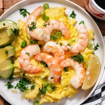 Shrimp and scrambled eggs topped with scallions and parsley, served with avocado slices and a lemon wedge on a white plate