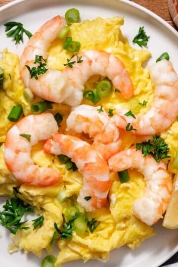 Shrimp and scrambled eggs topped with scallions and parsley, served with avocado slices and a lemon wedge on a white plate