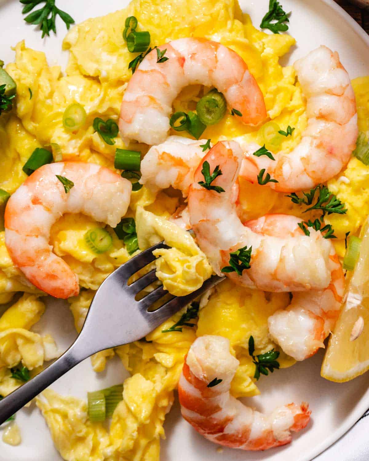 Shrimp and scrambled eggs with a fork lifting a bite, topped with scallions and parsley and served with a lemon wedge