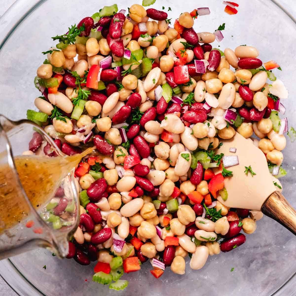 pouring vinaigrette over 3 bean salad with chickpeas, kidney beans, white beans, and vegetables in a bowl
