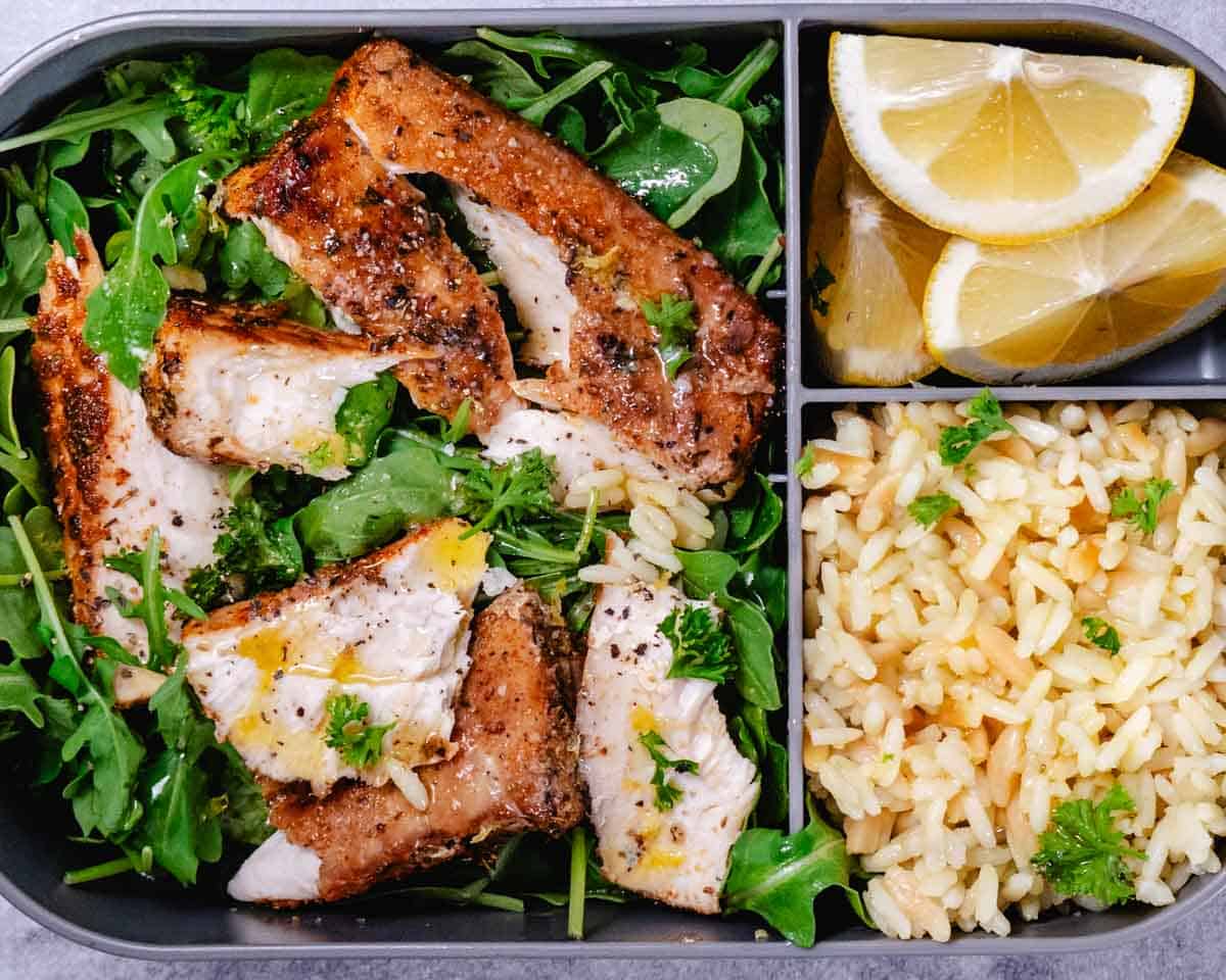 Blackened mahi mahi meal prep bowl with rice, mixed greens, and lemon wedges in a black container