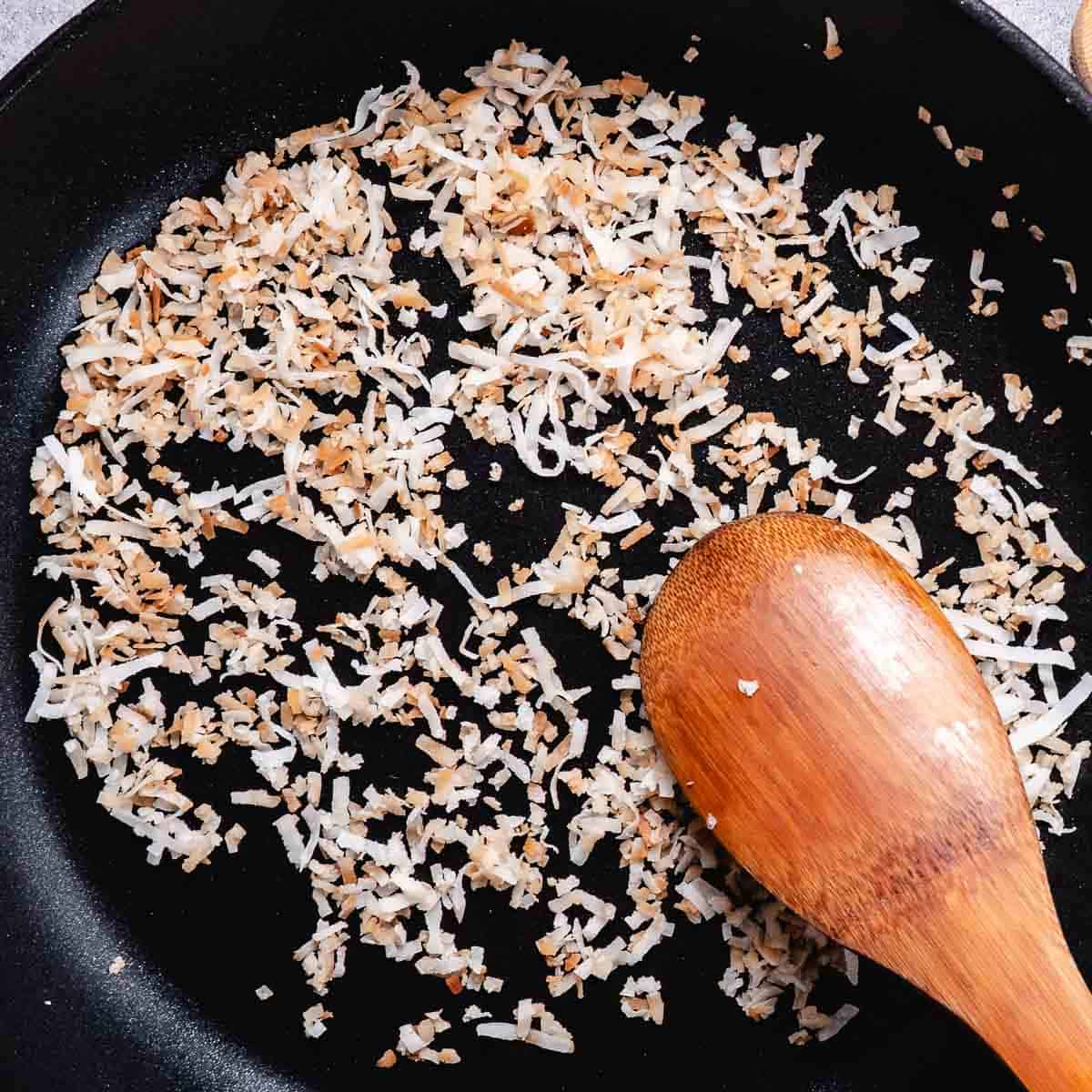 Toasted coconut in a black skillet with a wooden spoon.