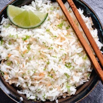 Coconut rice with lime wedges, lime zest, toasted coconut, and chopsticks in a dark bowl.