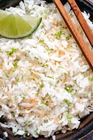 Coconut rice with lime wedges, lime zest, toasted coconut, and chopsticks in a dark bowl.