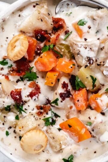 Creamy fish chowder with cod, potatoes, carrots, bacon, parsley, and oyster crackers in a white soup bowl.
