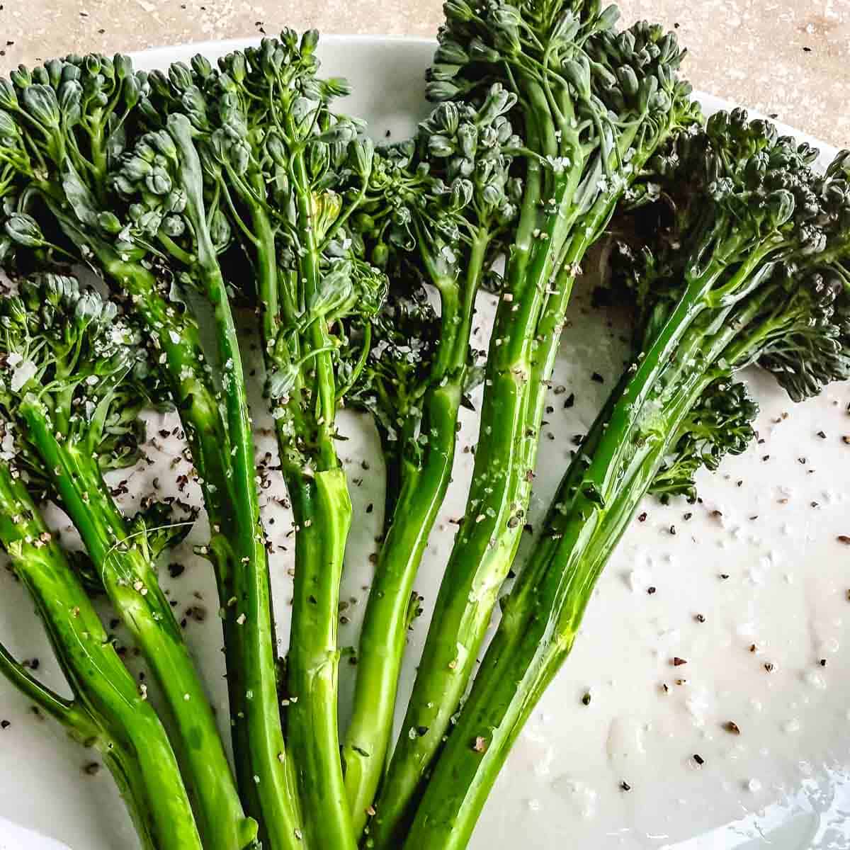 Raw broccolini coated with olive oil, coarse salt, black pepper, and herbs on a white plate before grilling.
