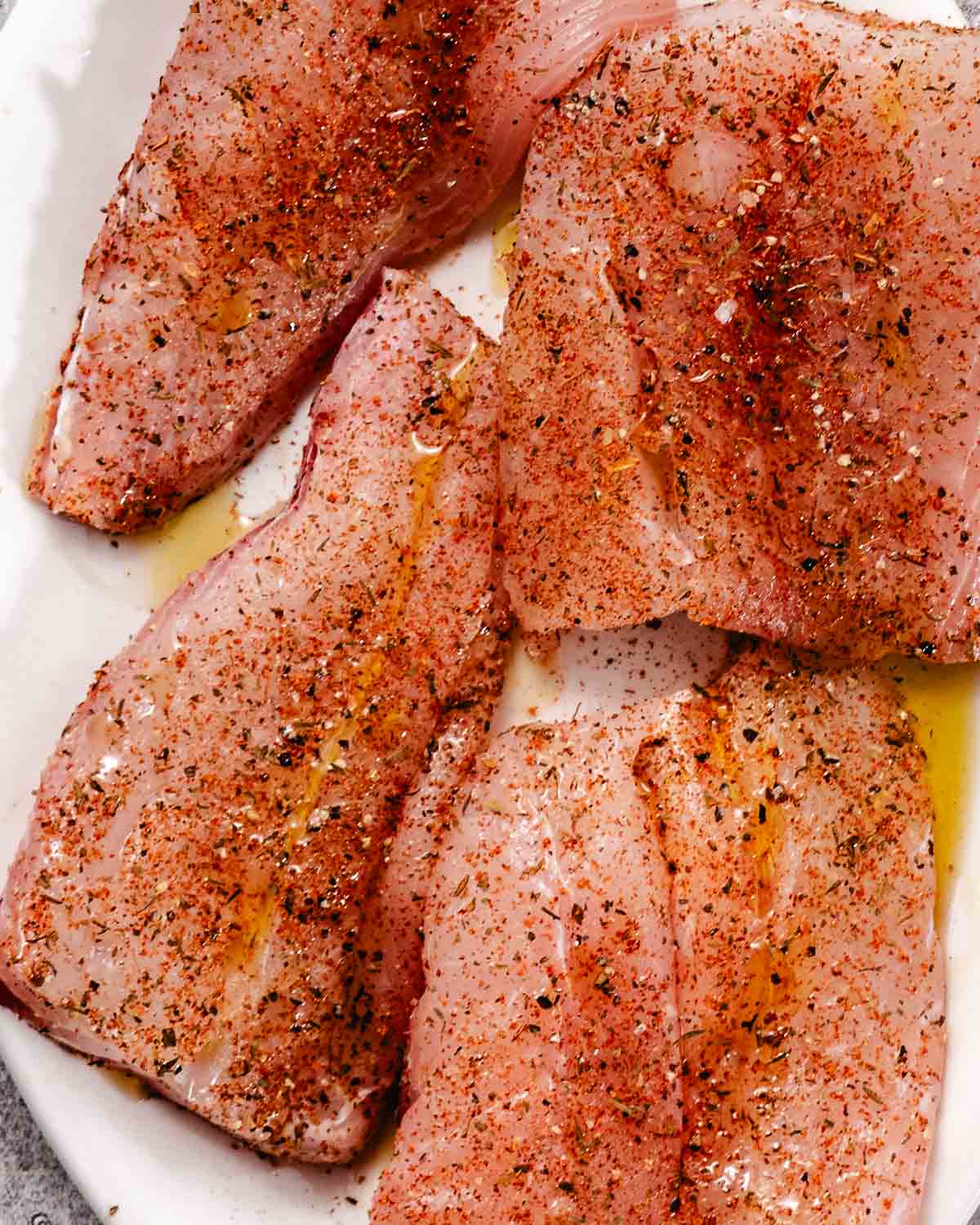 Raw red snapper fillets coated with olive oil and blackened seasoning on a white plate before grilling