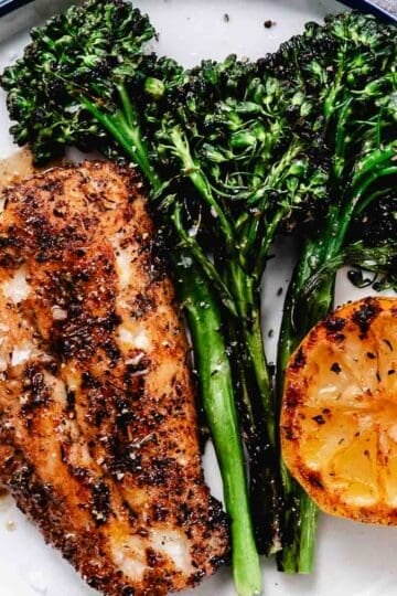Grilled red snapper fillet with charred baby broccoli and grilled lemon on a white plate with blackened seafood seasoning