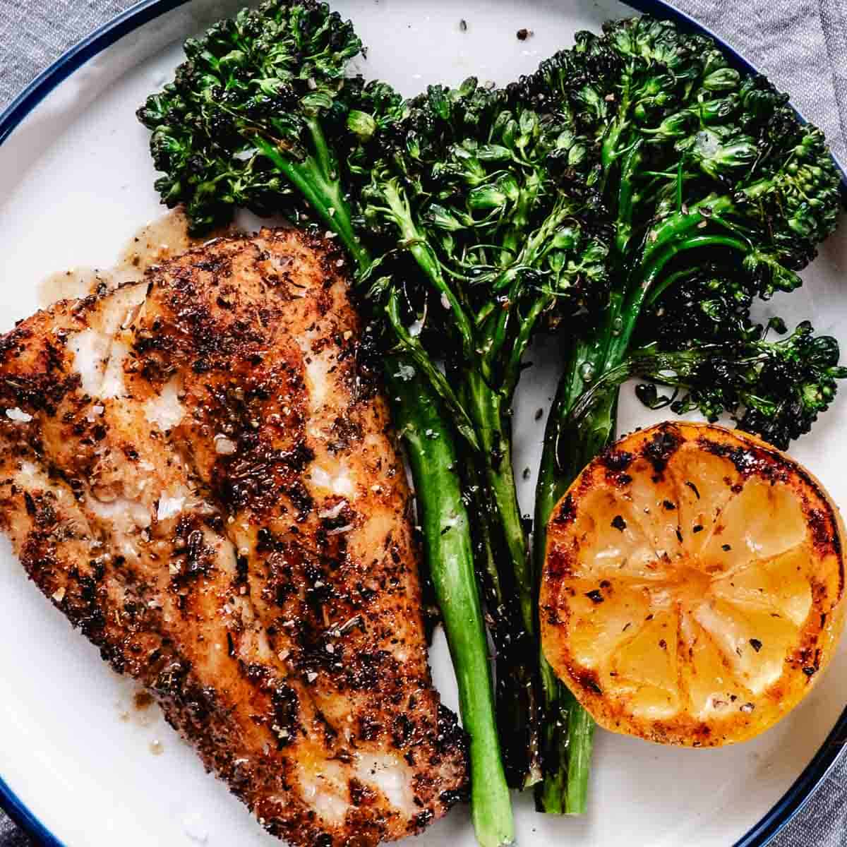 Grilled red snapper fillet with charred baby broccoli and grilled lemon on a white plate with blackened seafood seasoning