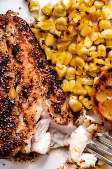 Grilled red snapper fillet with charred corn and grilled lemon on a white plate with flaky white fish pulled apart by a fork
