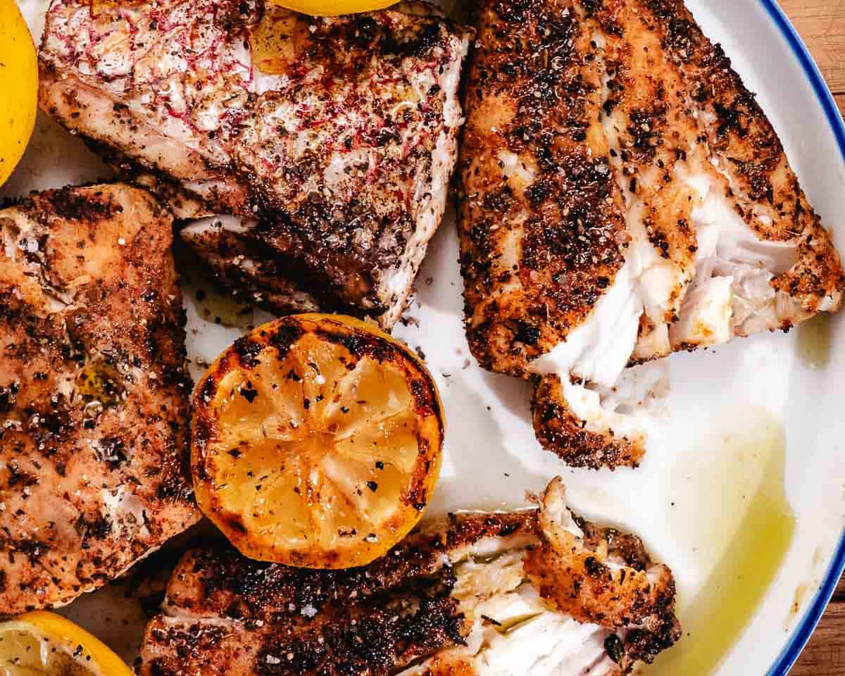 Plate of grilled red snapper fillets with charred lemons, flaky white fish, olive oil, and seafood seasoning on a white serving platter
