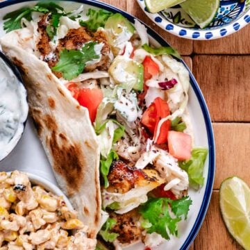 Grilled red snapper tacos with cabbage slaw, avocado, cilantro, and creamy sauce on toasted flour tortillas, served with Mexican street corn salad and lime wedges on a white plate.