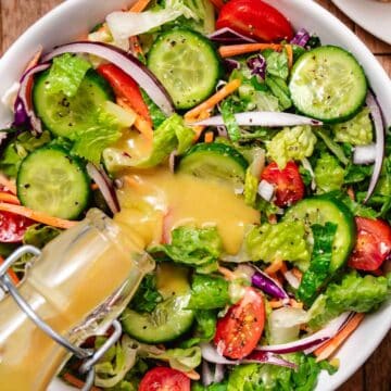 Creamy honey mustard dressing being poured over a fresh salad with romaine lettuce, cucumbers, cherry tomatoes, carrots, and red onion in a big bowl