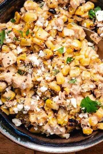 Mexican street corn salad with grilled corn, creamy dressing, cotija cheese, and fresh cilantro served in a black bowl with a serving spoon.