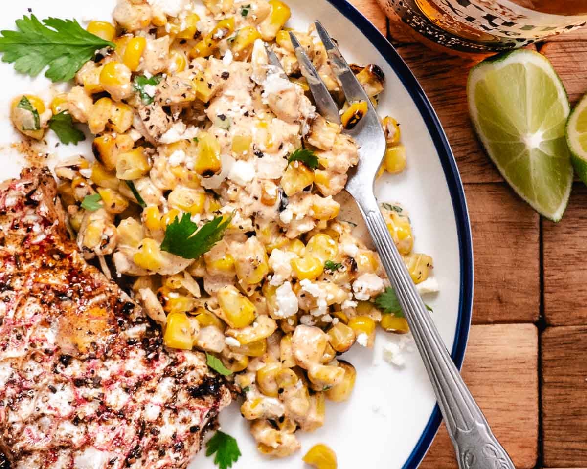 Mexican street corn salad served with grilled steak, lime wedges, and crumbled cotija cheese on a white plate. Creamy elote-style corn salad with charred grilled corn and fresh cilantro.